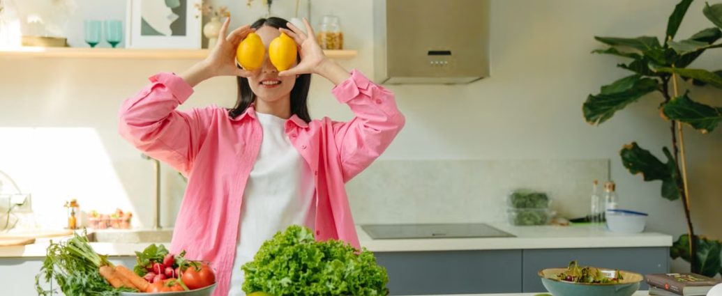 A woman playfully holding lemons over her eyes in a bright, lively kitchen setting filled with fresh vegetables.